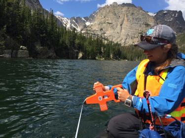 Isabella Oleksy takes depth measurements from an inflatable boat on a subalpine lake with large cliffs overhead