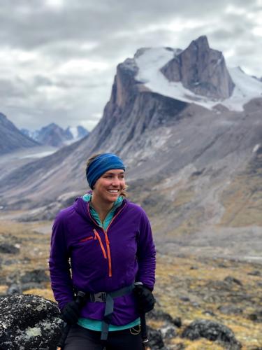 Sarah Crump, in purple coat, stands in front of a dramatic Baffin Island landscape of colorful tundra suddenly broken by towering cliffs.