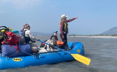 Irina Overeem standing and pointing while in a bright blue raft with Suzanne Anderson and others while studying river erosion in Alaska.