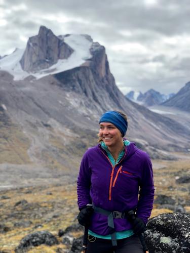 Sarah Crump, in purple coat, stands in front of a dramatic Baffin Island landscape of colorful tundra suddenly broken by towering cliffs.