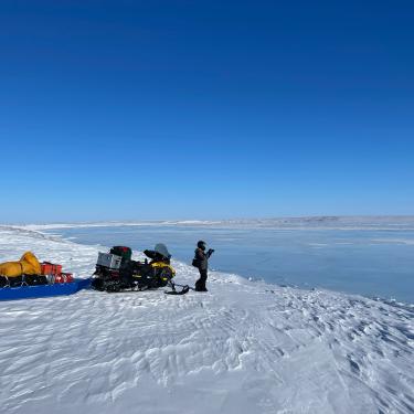 A snowmobile and an even larger attached sled are loaded with gear and parked on a snowy high river bank of the Canning River in Alaska. Standing next to them is Irina Overeem, who is scoping out how to cross the frozen river to a field site.