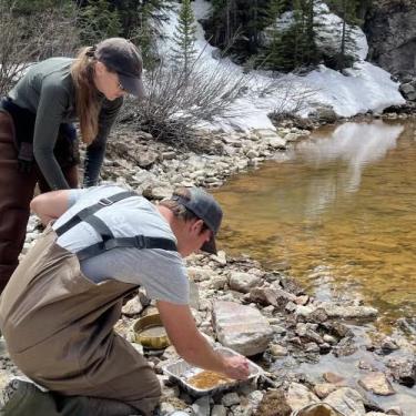 Two scientists collect water samples from a mountain stream, with melting snow on some of the snowbanks