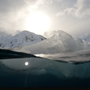 Ice floats at the surface of a cold ocean. The camera is half-submerged, showing both above and under water.