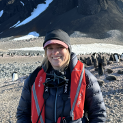 Cassandra Brooks, in warm puffy jacket and brimmed cap, stands near a penguin colony with a dark cliff behind