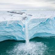 A stream on top of an ice shelf ends abruptly in a dramatic waterfall, splashing into the sea.  Photo by Florian Ledoux of the Arctic Arts Project