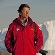 James Balog, in a red jacket with a long-lens camera slung over his shoulder, looks into the distance of an icy landscape.  Behind him is a large jagged iceberg.