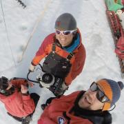Overhead view of three smiling Antarctic researchers wearing red coats, hats, and sunglasses.  Michael Dyonisius grips a powered ice auger with two hands.  Right next to him are Sarah Shackleton and Bernhard Bereiter.  Photo taken in austral summer of 2015-2016.  Photo overlain with text saying: Meet Michael Dyonisius, INSTAAR postdoc and expert in greenhouse gases, radiocarbon, and ice cores.