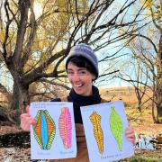 On a fall day, Lauren Magliozzi, in knit hat and overalls, holds up large drawings of common diatoms of Coal Creek in Colorado