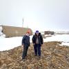 Two men in winter gear stand on a bare patch of tundra with a snowy research station in the background
