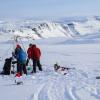 Researchers stand around a complex of beams, cords and instruments next to an ice augur on a snowy lake with snow-covered rocky terrain in the background
