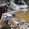 Two scientists collect water samples from a mountain stream, with melting snow on some of the snowbanks
