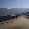 People in the foreground and background walk on very large sand dunes with arid mountains in the distance