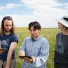Four young people in outdoor clothing look at a phone amidst a sun-lit meadow