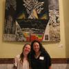 Jocelyn Catterson and Holly Barnard stand in front of Catterson's large painting on display in the Colorado capitol building rotunda. They are smiling and have their arms around each others' shoulders.