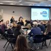 INSTAAR celebration luncheon May 8th 2024: A large room full of round tables packed with dozens of seated people having lunch and listening to opening remarks from Interim Director Nikki Lovenduski and Assistant Director of JEDI Julio Sepúlveda