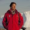 James Balog, in a red jacket with a long-lens camera slung over his shoulder, looks into the distance of an icy landscape.  Behind him is a large jagged iceberg.