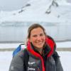 Cassandra Brooks, in a warm puffy jacket, stands near the shore of cold ocean waters with glacier peaks behind