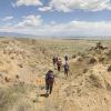 A group of paleontologists, wearing backpacks and sun hats, walk down a gully of eroding sedimentary rocks with a open vista of grassy plains in front of them. Photo by Rick Wicker DMNS