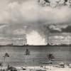 Black and white photo of a nuclear bomb test's mushroom cloud with nearby ships and palm trees in the foreground