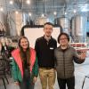 Emily Stuchiner, Lennart van Maldegem, and Michael Dyonisius pose near the small stage and screen used for INSTAAR's first storytelling night at Sanitas Brewing Company. In the background are big metal brewing tanks and a wall of wooden barrels.
