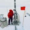 Glaciologist Tobias Erhardt, in puffy red parka and black pants, uses a big metal drill to extract a shallow ice core at the East Greenland Ice-core Project camp.