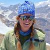 Millie Spencer stands on the sunny summit of Cerro El Plomo (17,795 feet) in Central Chile. Behind the rocky summit is a prominent glacier, with a background of mountain peaks with more glaciers and snowfields. She is wearing a green jacket, blue knit hat, and mirrored glacier glasses