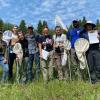 Nine researchers stand close together facing the camera, smiling and holding butterfly nets, clipboards, and water bottles. They stand in a grassy clearing surrounded by trees, under a blue sky.