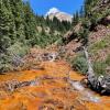A mountain stream in the upper Mancos river basin, displaying a rusty red bed, flows toward the viewer. Mountain slopes covered with pine trees are in the background. Photo by Andrew Manning.