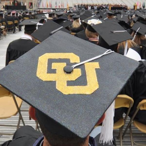 Graduation cap with gold CU letters and other graduates can be seen sitting in the background at the ceremony