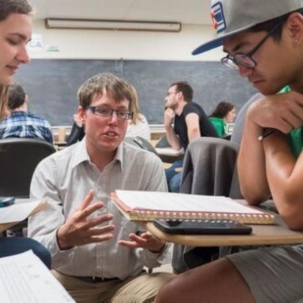 Professor in classroom squatting down and speaking to two students at their desks