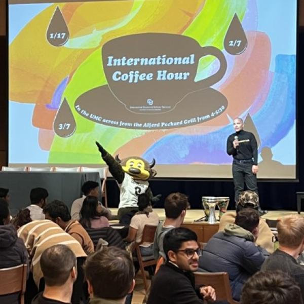 Chip posing in front of a slide presented at iternational student orientation with tables of students watching the presentation