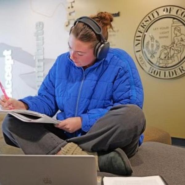 Person wearing headphones with a laptop writing in a notebook and studying for finals