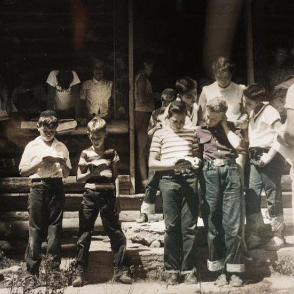 Black and white photo of a group of children camp-goers reading from their prayer books. In the right corner, a man in all white appears to be leading everyone.