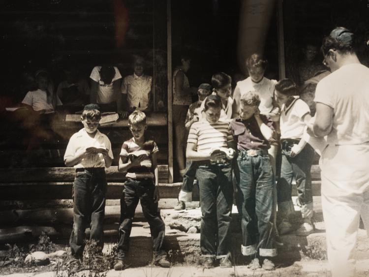 Black and white photo of a group of children camp-goers reading from their prayer books. In the right corner, a man in all white appears to be leading everyone.