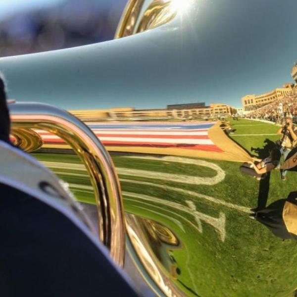 A person holding a brass marching band instrument with a reflection of the CU football field during CU vs. Stanford in 2019.