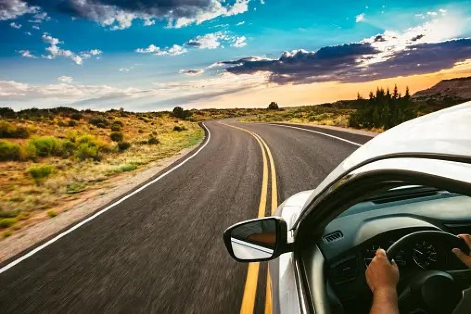 A person driving in a silver car on an open highway with desert landscape and blue sky with clouds in the horizon.