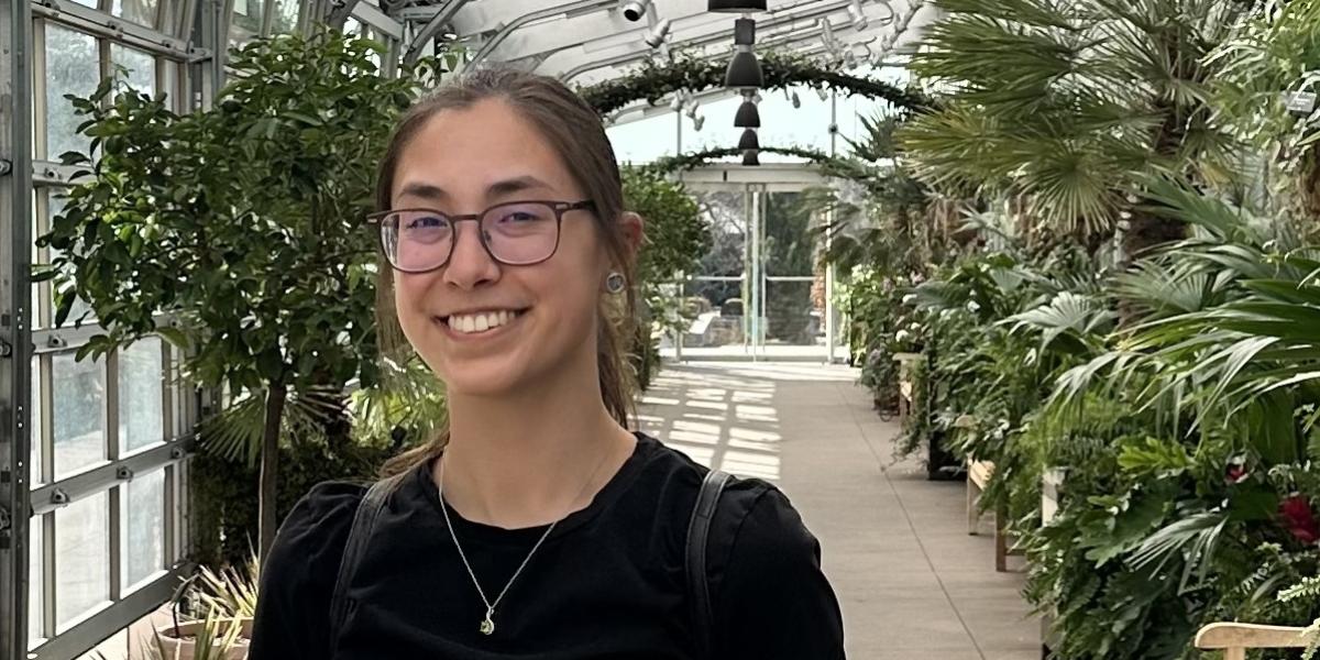 Jenna smiling wearing glasses and a black shirt in front of some trees in a green house.