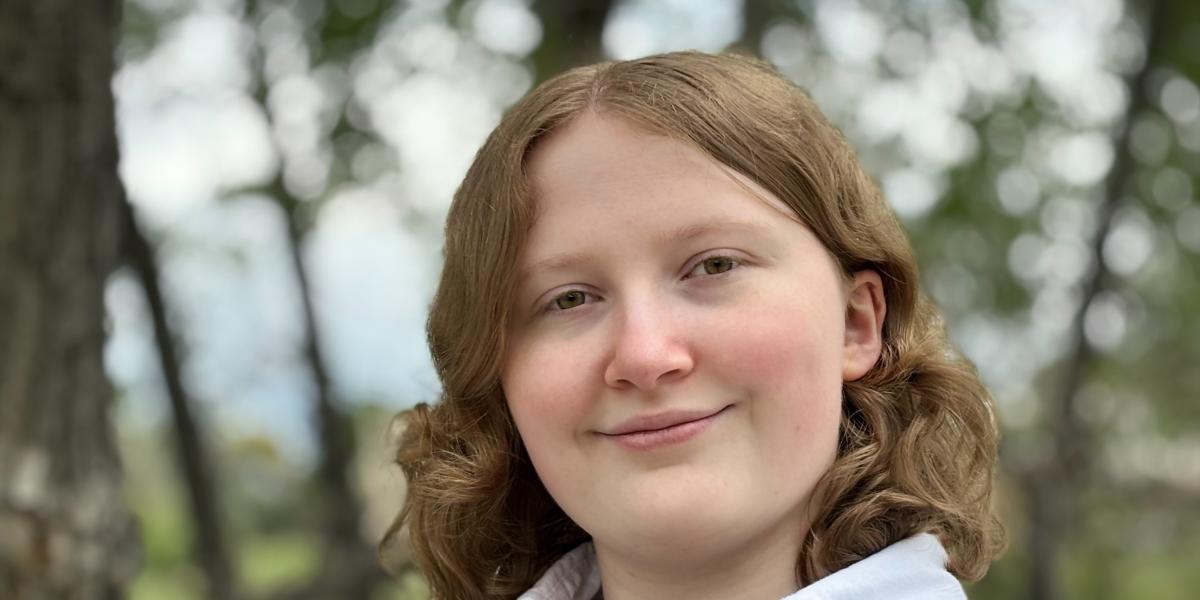 Picture of Kelly smiling in a white collared shirt with a forrest in the background