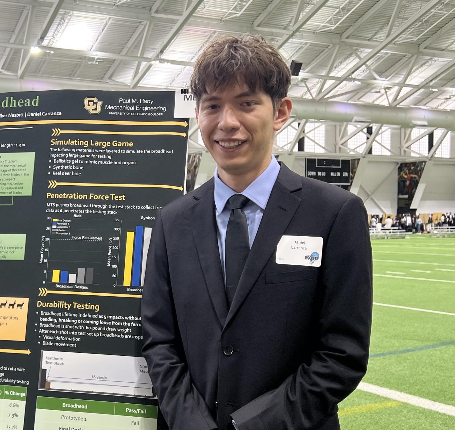 Daniel standing at Senior Design Expo in front of poster & field in suit.