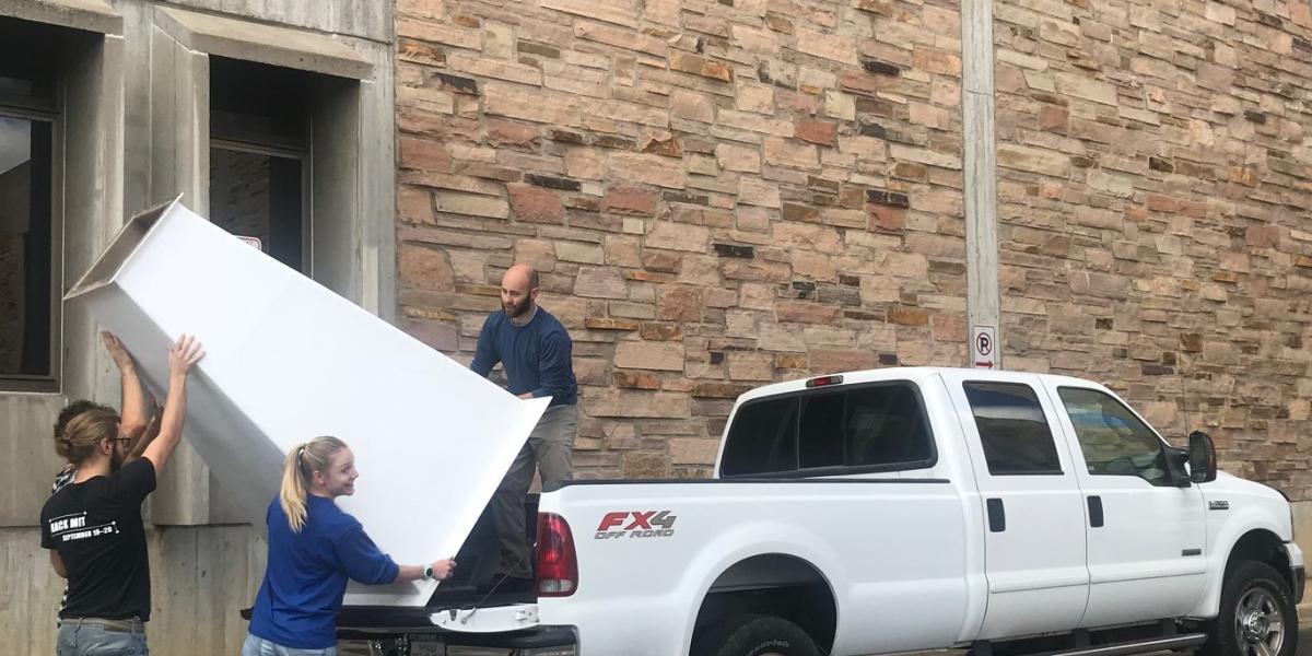 A couple of students heaving a large piece of equipment onto the back of a truck