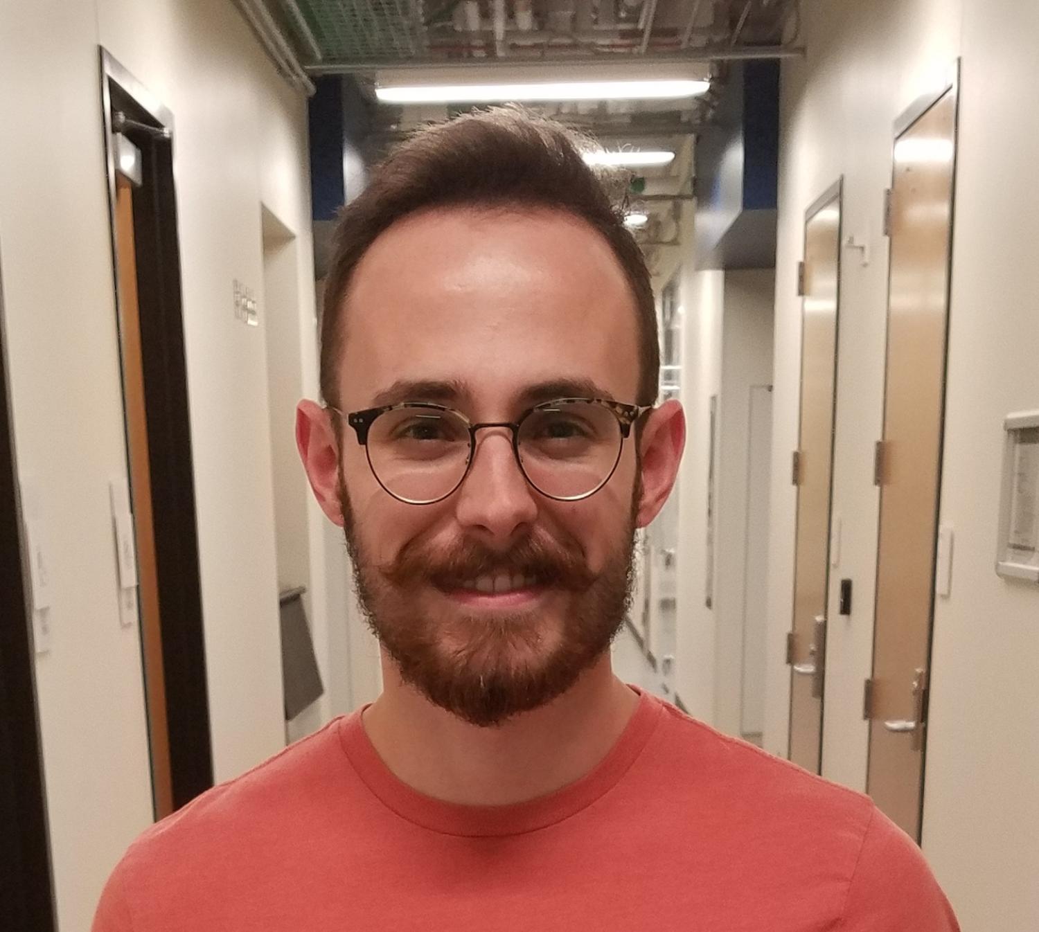 Evan Bisirri. He is smiling and wearing an orange shirt in the middle of a hallway.