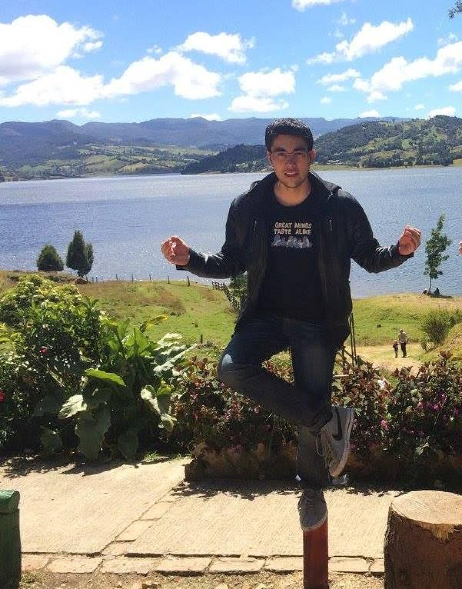 Andres Chaparro Sosa. He is balancing on one foot on a pole. There is a lake and mountains behind him.