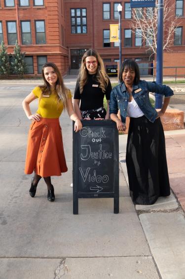 Three people stand around a sign that says, "Check out Justice by Video," a conference given by Sandra Rivstovska.