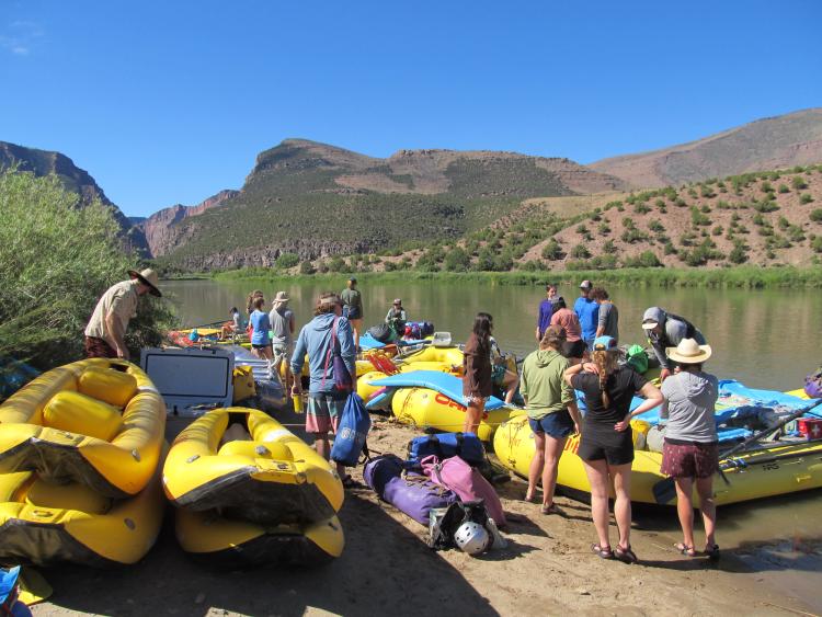 Yellow river rafts pulled on the shore of a river in the mountains with students gathered around.
