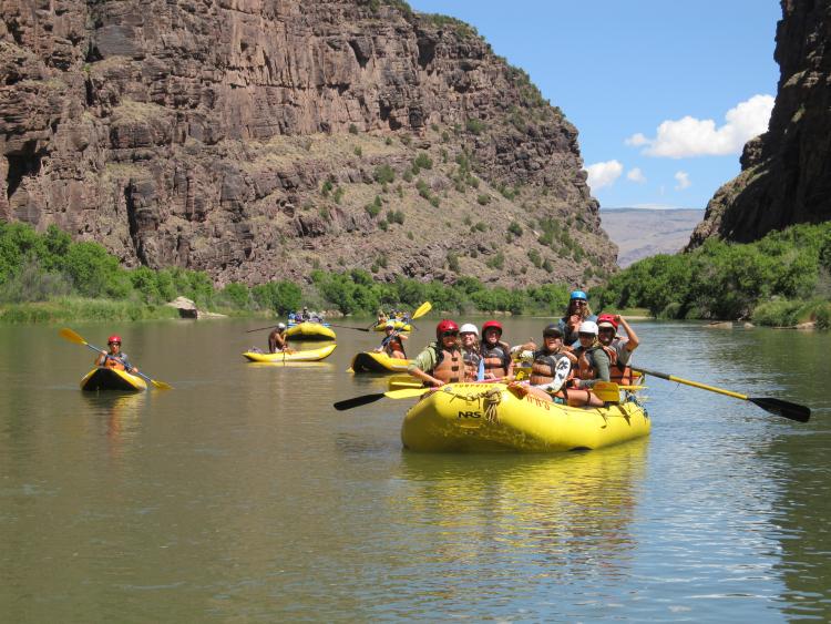 A group of students float on a yellow raft in the middle of a river.