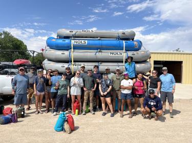 A group of students and NOLS facilitators stand in front of a trailer stacked high with river rafts