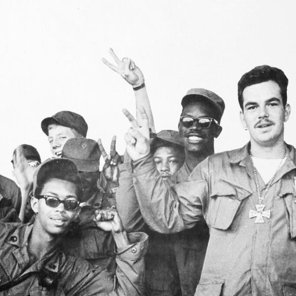 Ten young men in military garb smile at the camera, some flashing the peace sign