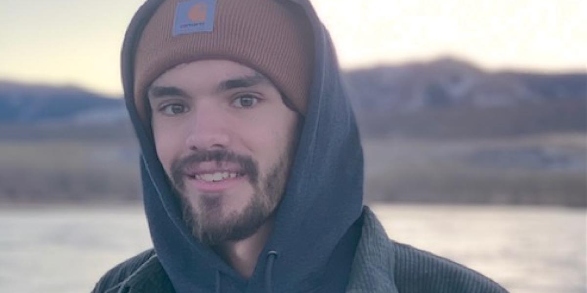 A person in a blue hoodie and a beanie stands in front of a mountain lake.