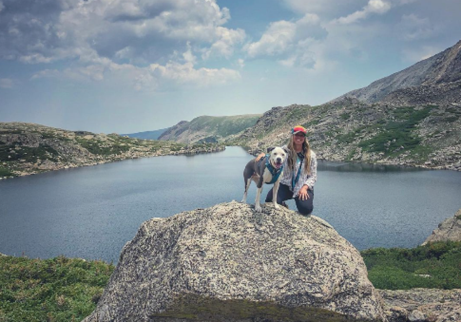 Dr. Rai Farrelly kneels on a large boulder next to a grey and white dog, with a mountain lake in the background and a cloud-strewn sky overhead