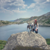 Dr. Rai Farrelly kneels on a large boulder next to a grey and white dog, with a mountain lake in the background and a cloud-strewn sky overhead
